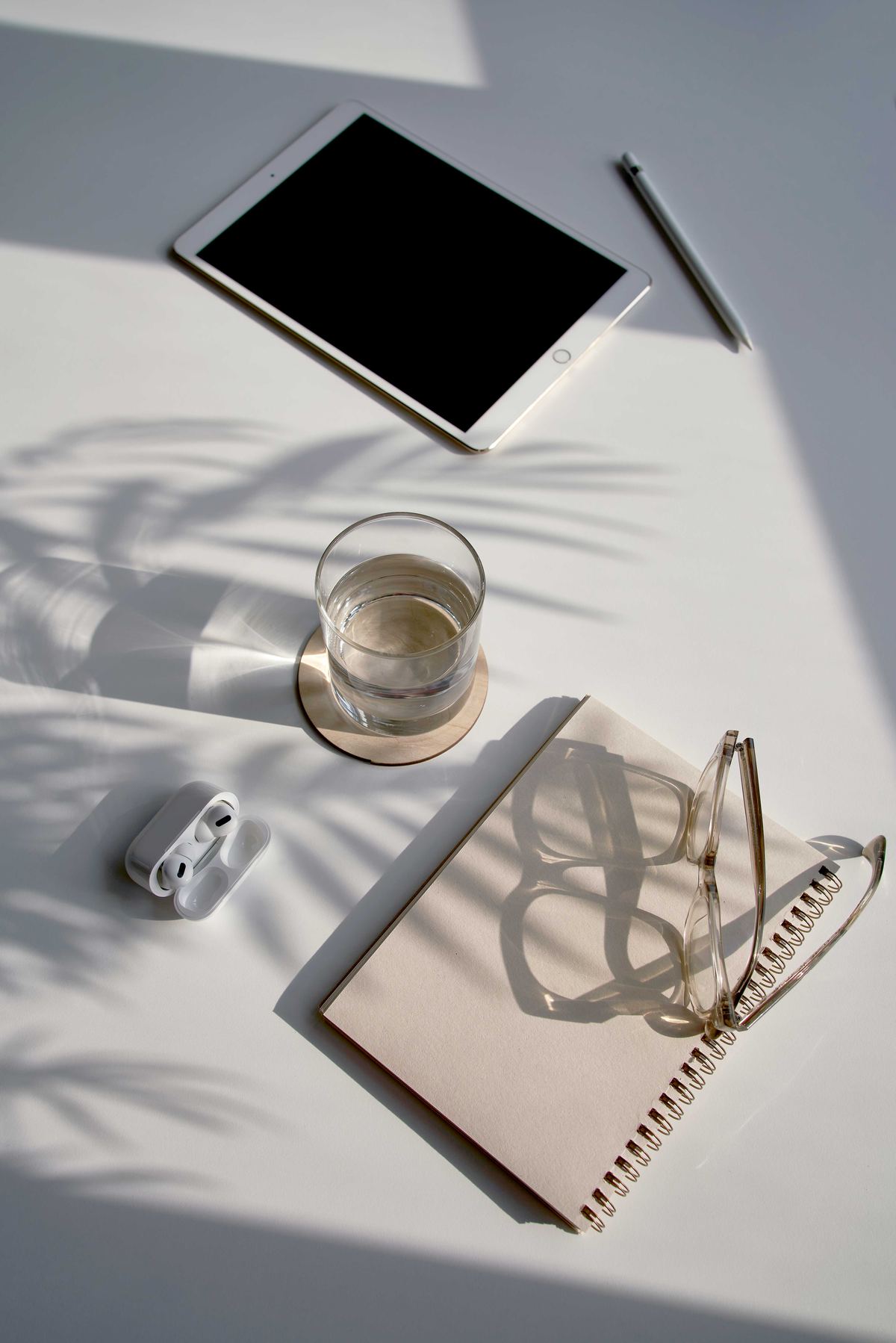 Cuaderno, tableta y gafas sobre una mesa blanca con sombras de plantas